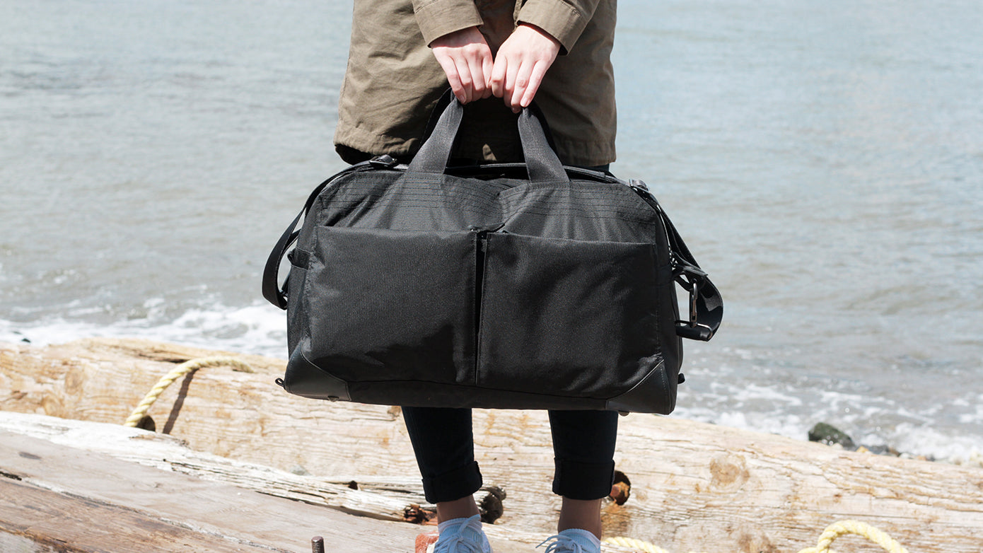 Person holding a black V1 Pakt One Duffel bag by the water's edge, standing on wooden logs.