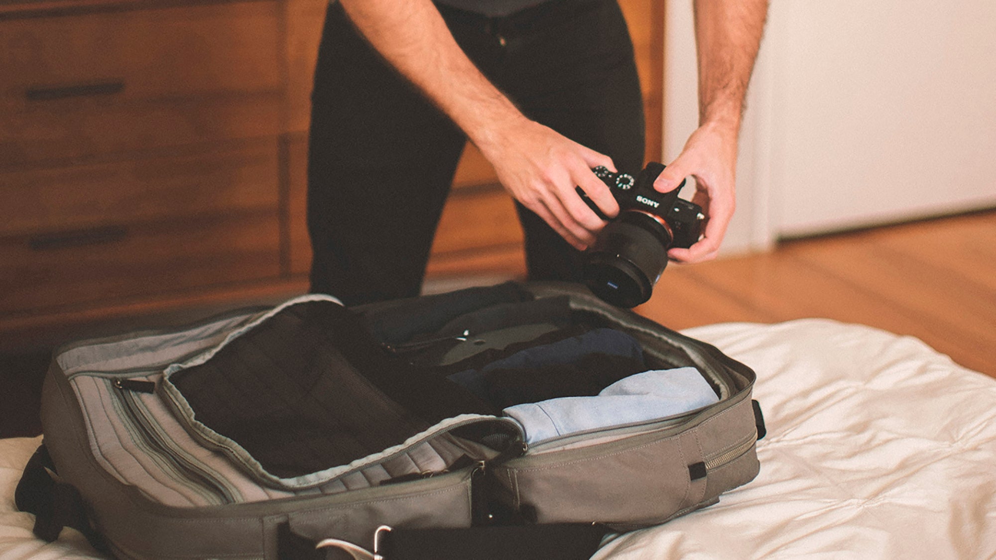 Person holding a Sony camera over an open Pakt One with folded clothes on a bed in a room