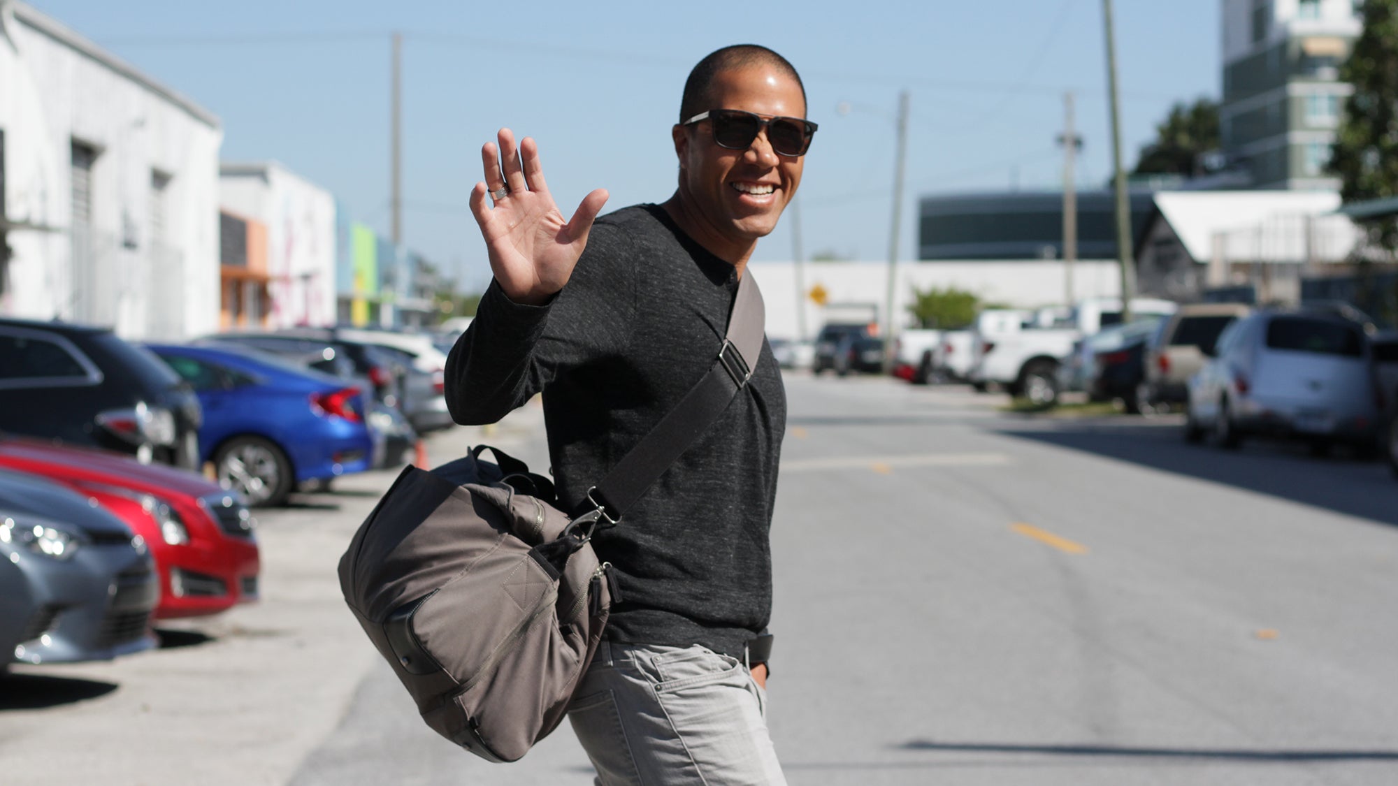 Smiling man wearing sunglasses waving with his right hand, carrying a gray V1 Pakt One bag, standing on a street with parked cars and buildings in the background.