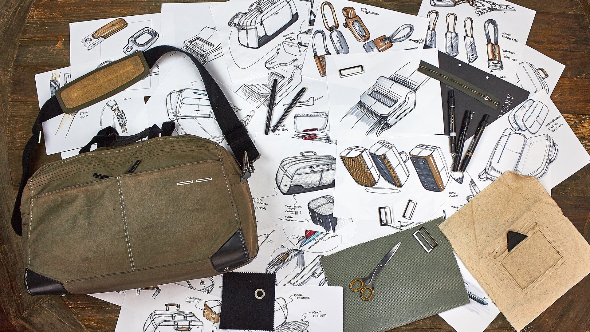 An early prototype on a wooden table surrounded by sketches of bags, fabric swatches, scissors, and pens.