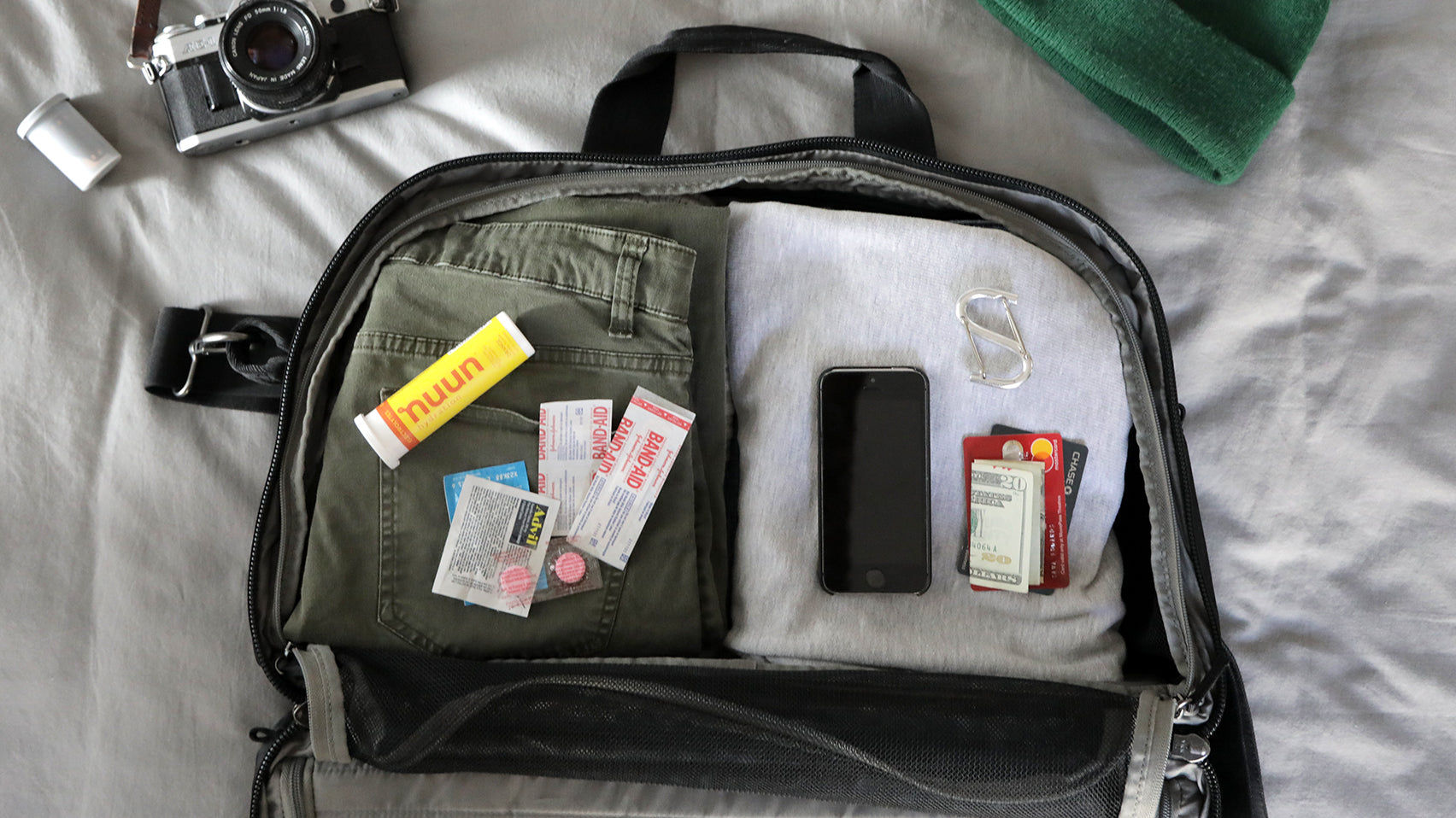 An open V1 Pakt One Travel Bag with folded green pants, gray shirt, smartphone, cash, credit cards, band-aids, medication, and a carabiner clip on a bed.