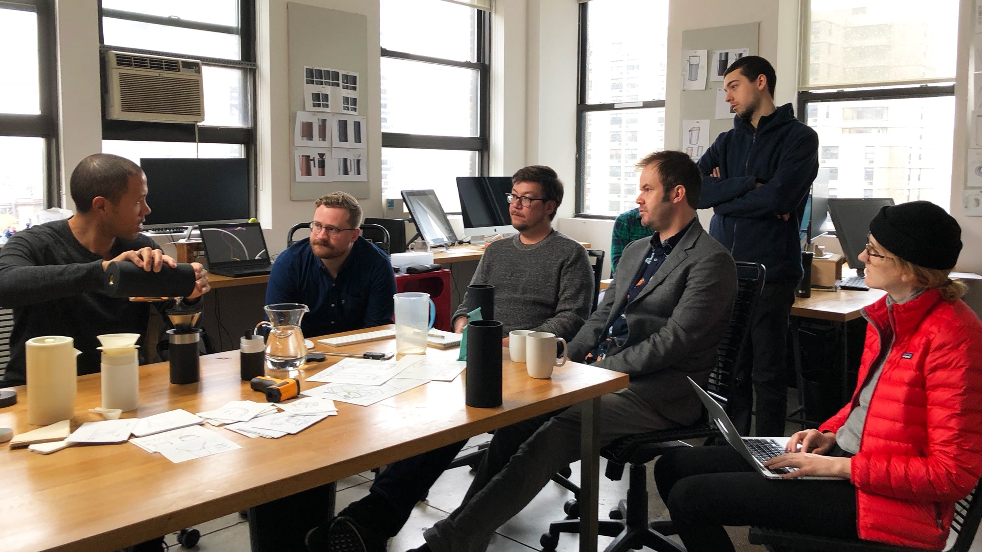 Six people in an office meeting around a table with sketches, mugs, and pitchers, discussing a product prototype.