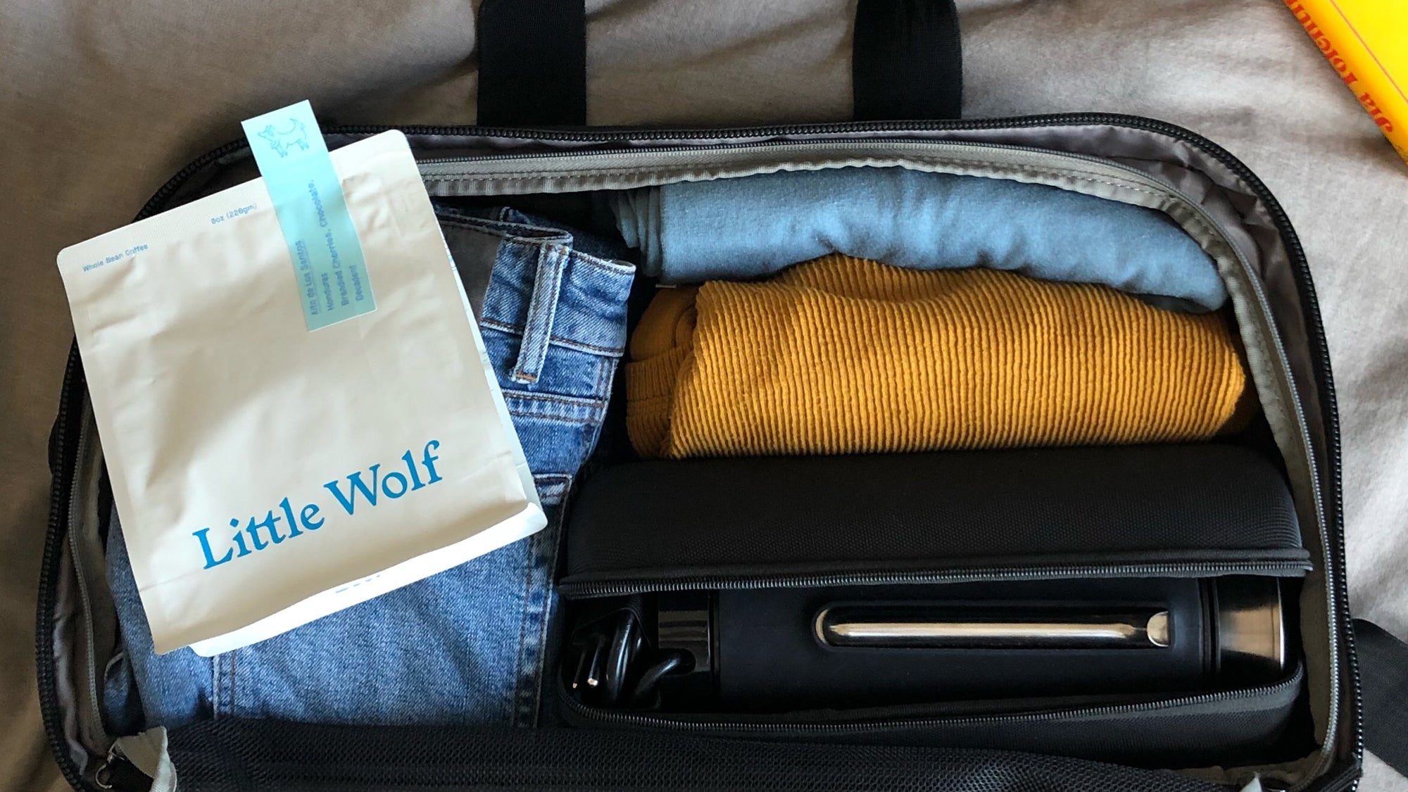 A packed V1 Pakt One with folded clothes and a white coffee bag labeled 'Little Wolf' with a blue sticker reading 'With the same roasting method, organic, chocolate, balanced'.