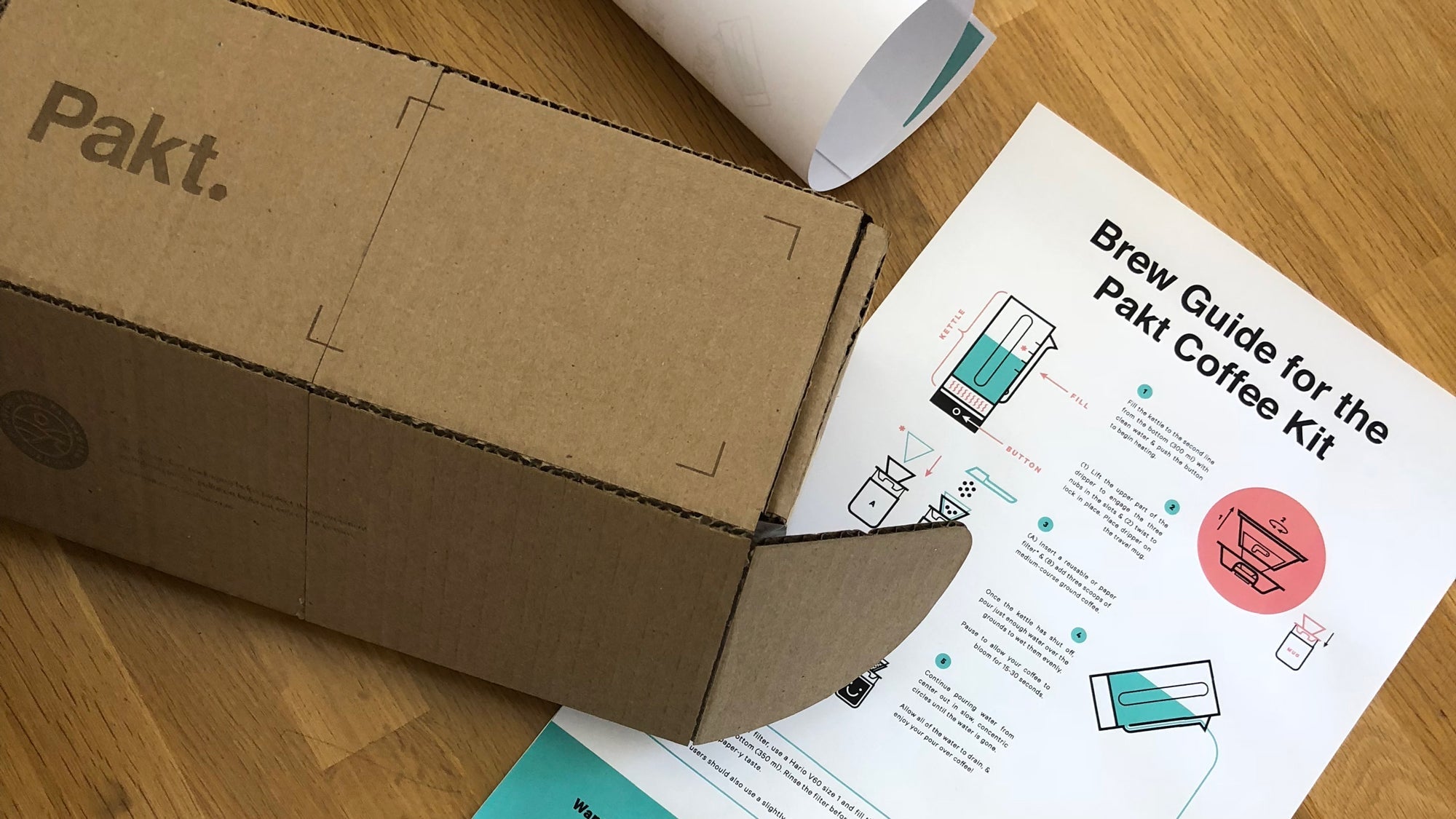 Cardboard box with 'Pakt.' logo and a Brew Guide for the Pakt Coffee Kit showing brewing instructions and illustrations on a wooden surface.