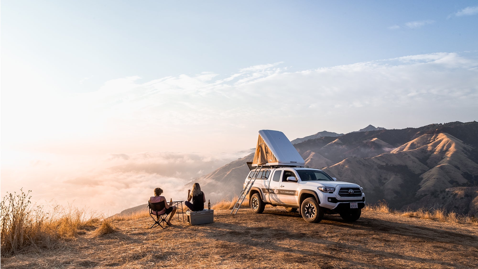 Two people sitting on camping chairs near a white camper truck with a rooftop tent on a mountain ridge at sunset, overlooking clouds and distant mountains.