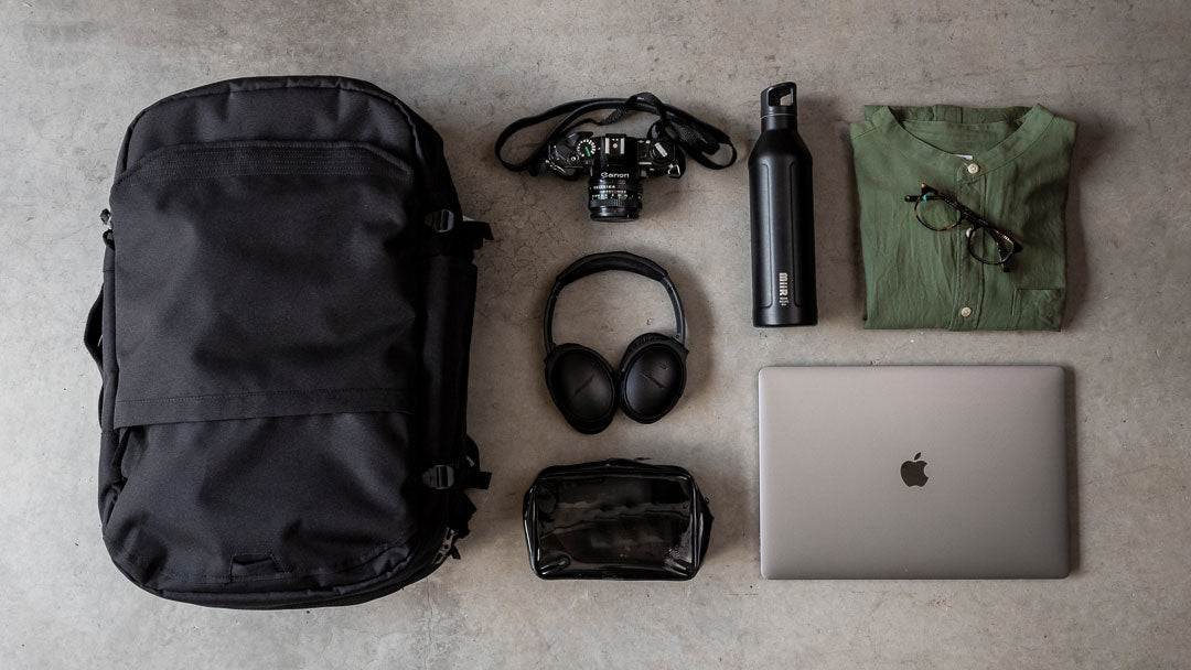 Black V1 Travel Backpack with a vintage camera, black headphones, black water bottle, folded green shirt with glasses, black toiletry bag, and a closed silver laptop arranged on a gray surface.