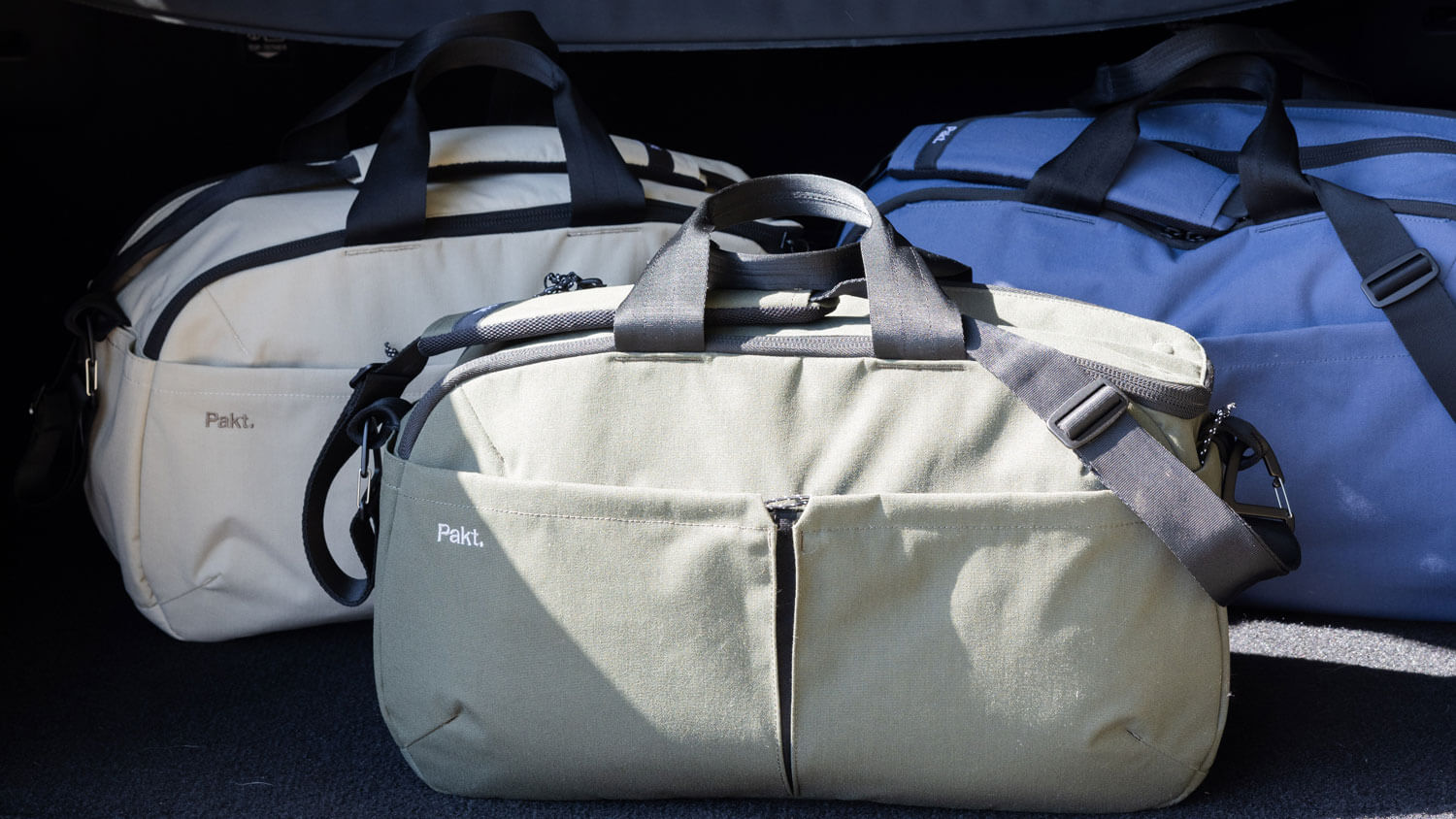 Three Pakt One Travel Duffels in Sand, Forest, and Ocean with black handles and shoulder straps, two bags have the brand name 'Pakt.' visible on the front pocket.