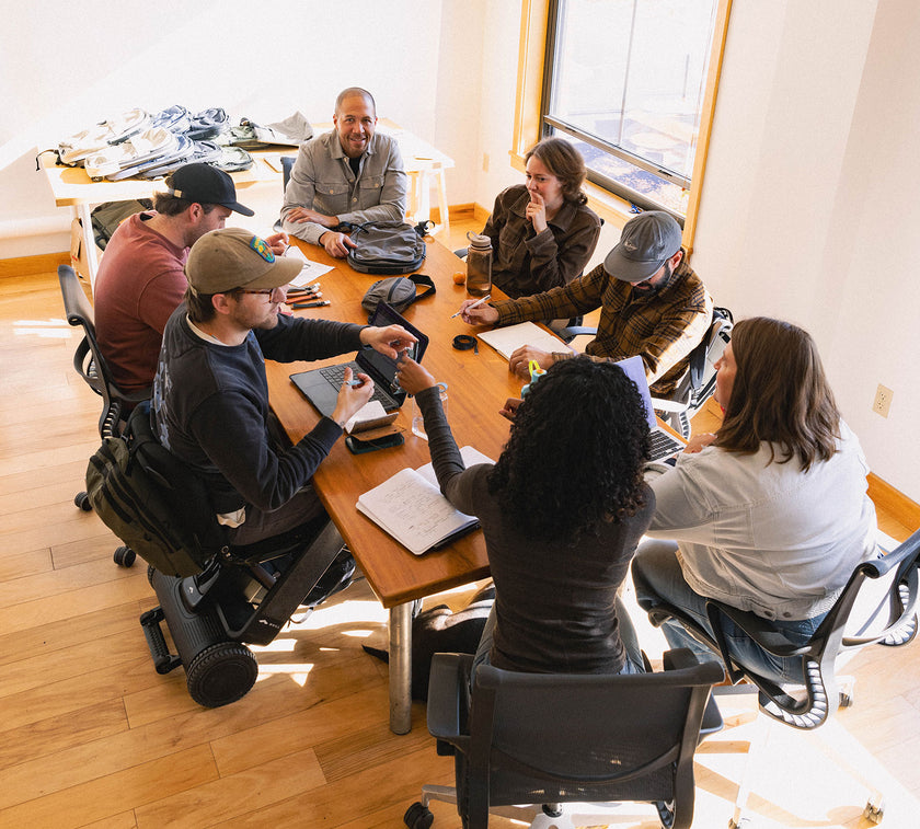 The Pakt team of seven people collaborating around a wooden table in a bright room, some using laptops and notebooks.