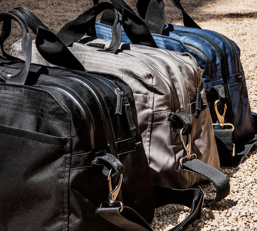 Three V1 Pakt Ones in black, gray, and blue with zippers and shoulder straps placed on gravel ground.