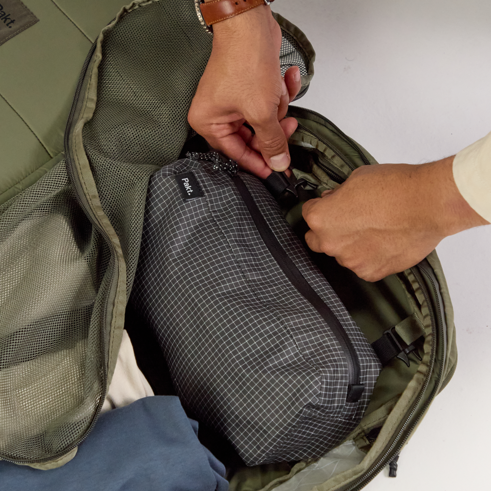 Hands placing a gray, grid-pattern packing cube labeled 'Pakt' into an open olive-green backpack.