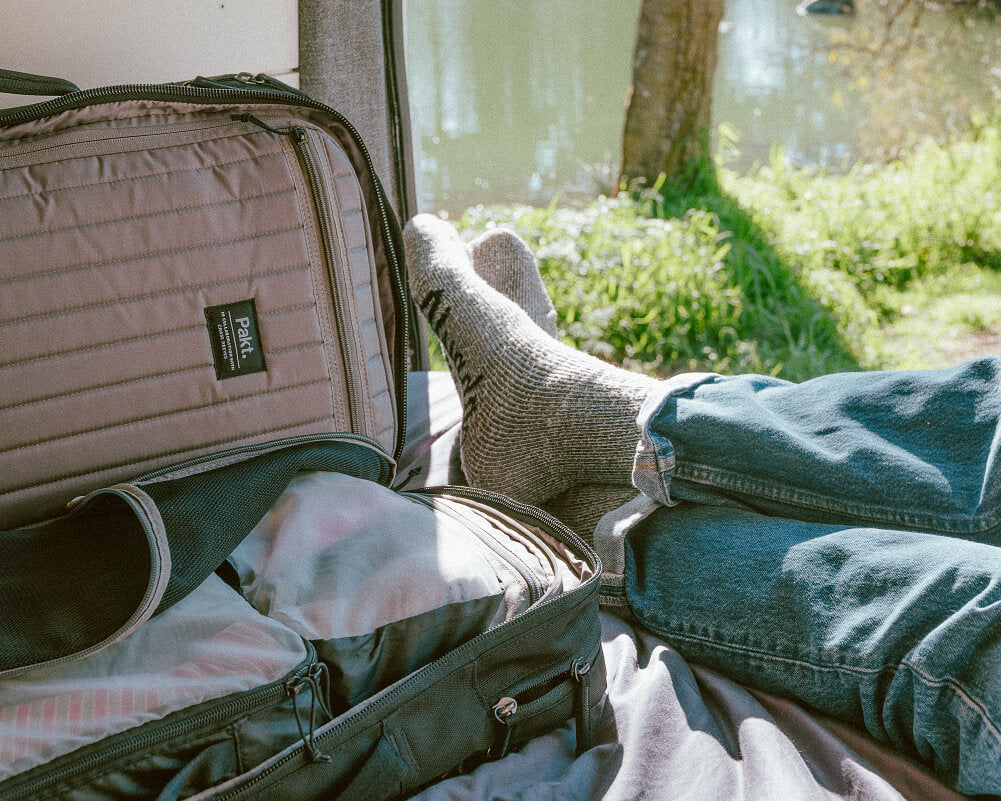 Open travel backpack with packed clothes beside legs in jeans and wool socks at a lakeside campsite; label 'Pakt.' visible