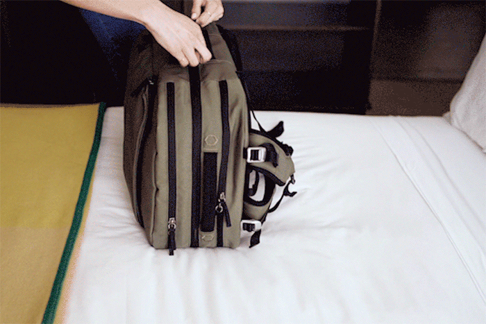 Hands zipping an olive-green backpack on a white bed.