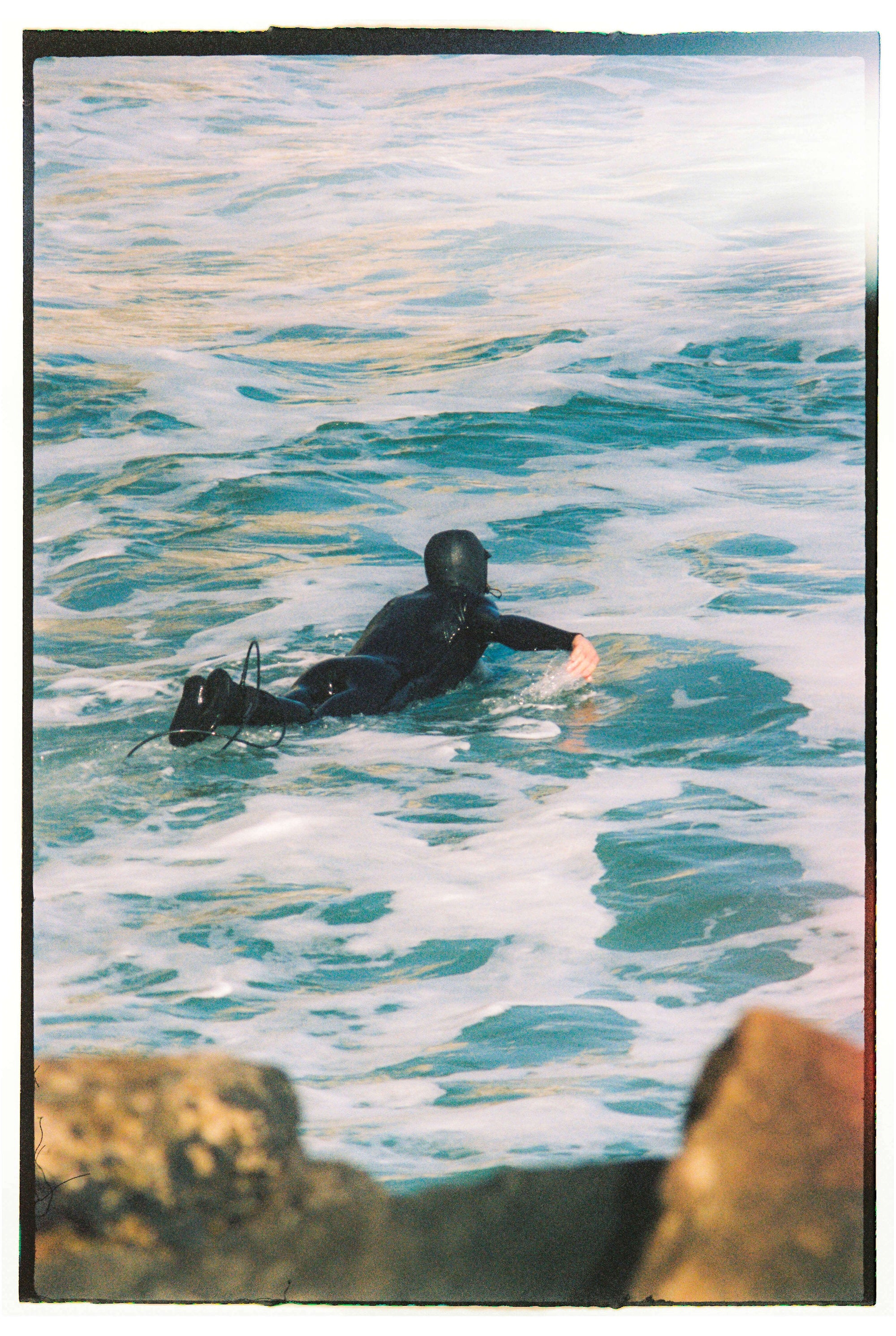Person in a black wetsuit paddling on ocean water with rocks in the foreground