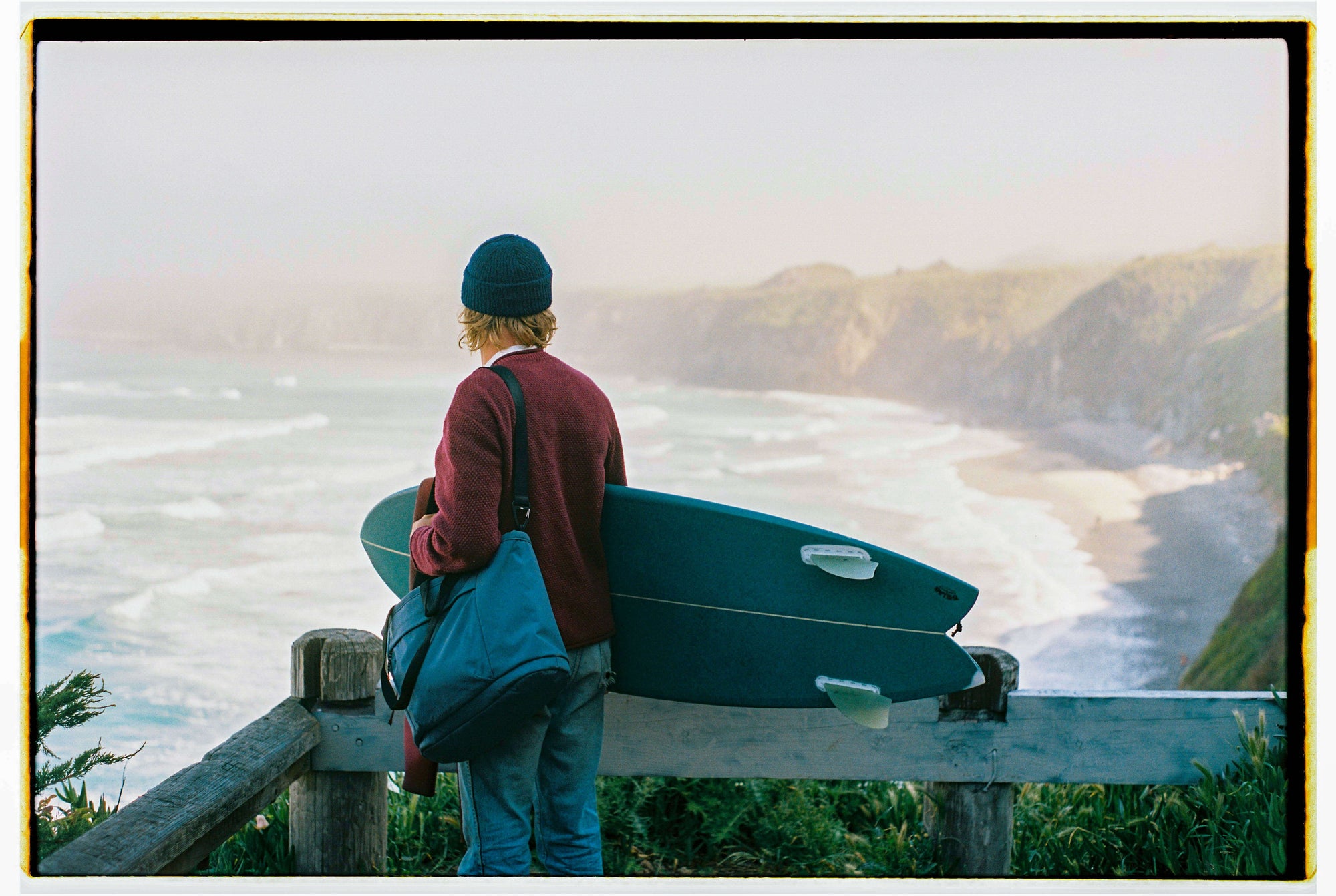 Person wearing a blue beanie and red sweater holding a blue surfboard and a blue bag, standing on a wooden fence overlooking a misty coastal cliff and ocean waves.