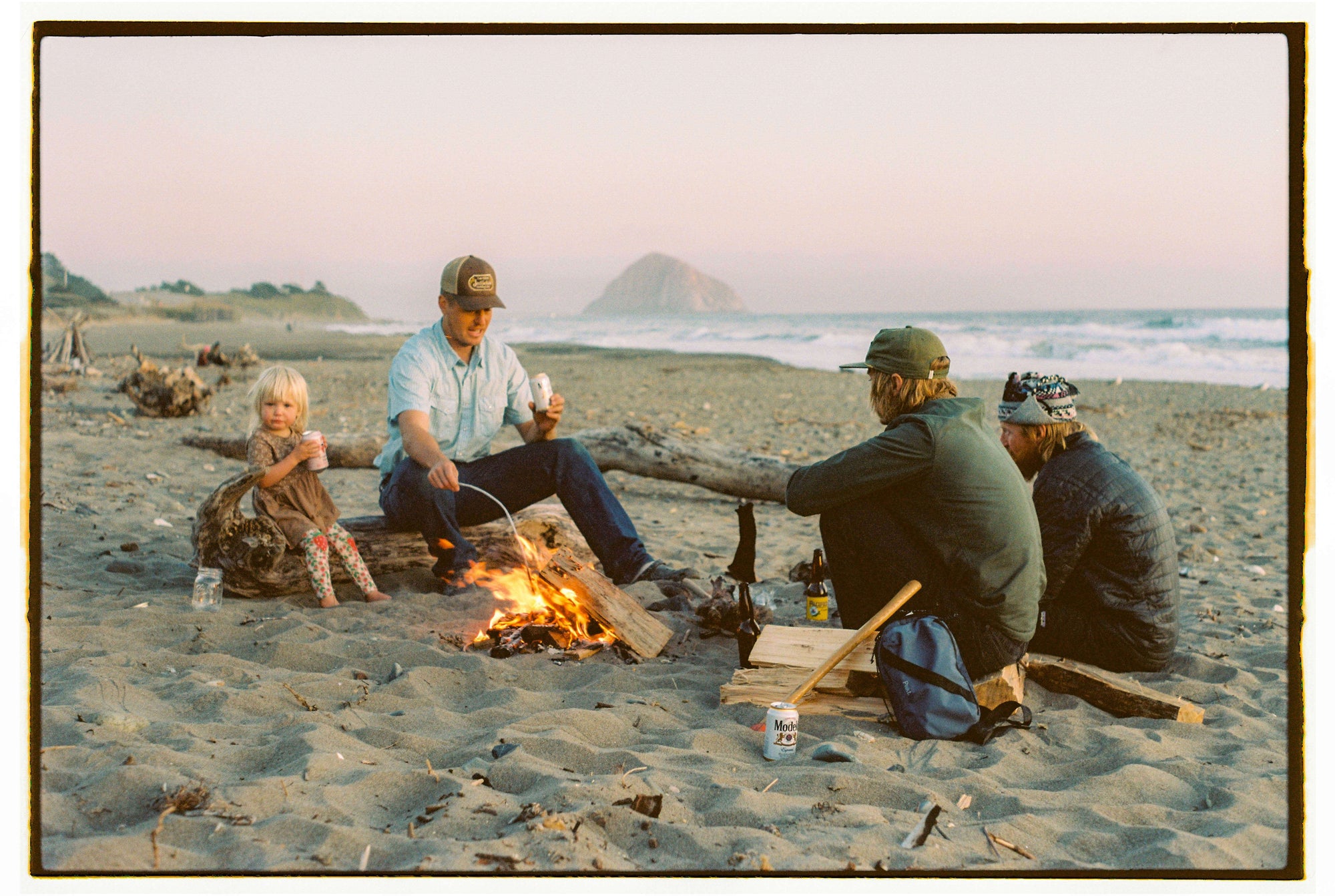 A group of three adults and one child sitting around a campfire on a sandy beach near the ocean at sunset.