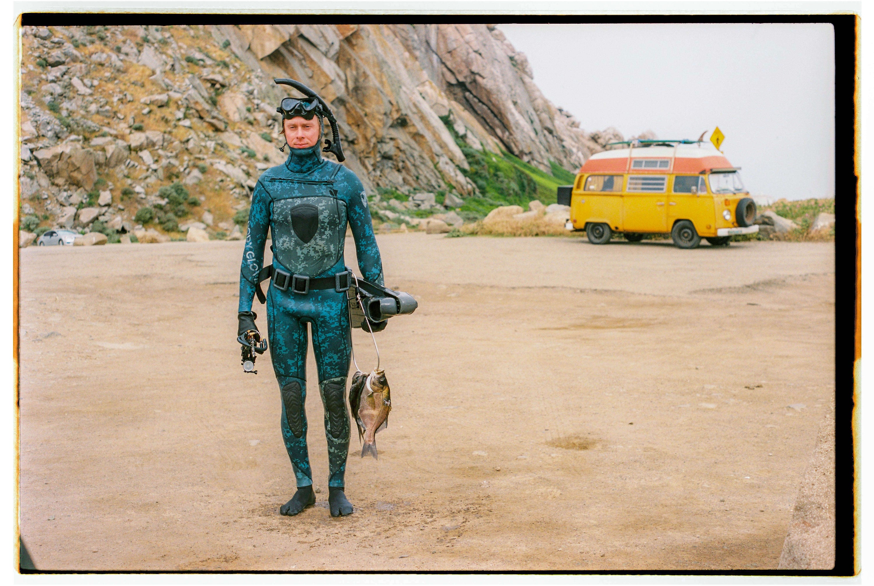 Person in a blue camouflage wetsuit holding two fish and diving equipment, standing on a dirt area with rocky cliffs and a yellow van in the background.
