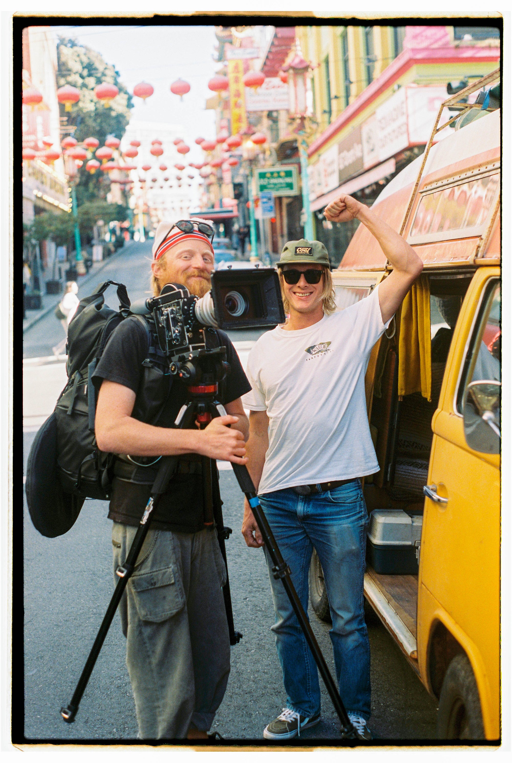 Two men on a street decorated with red lanterns; one holds a professional camera on a tripod, the other flexes his arm next to a yellow van.