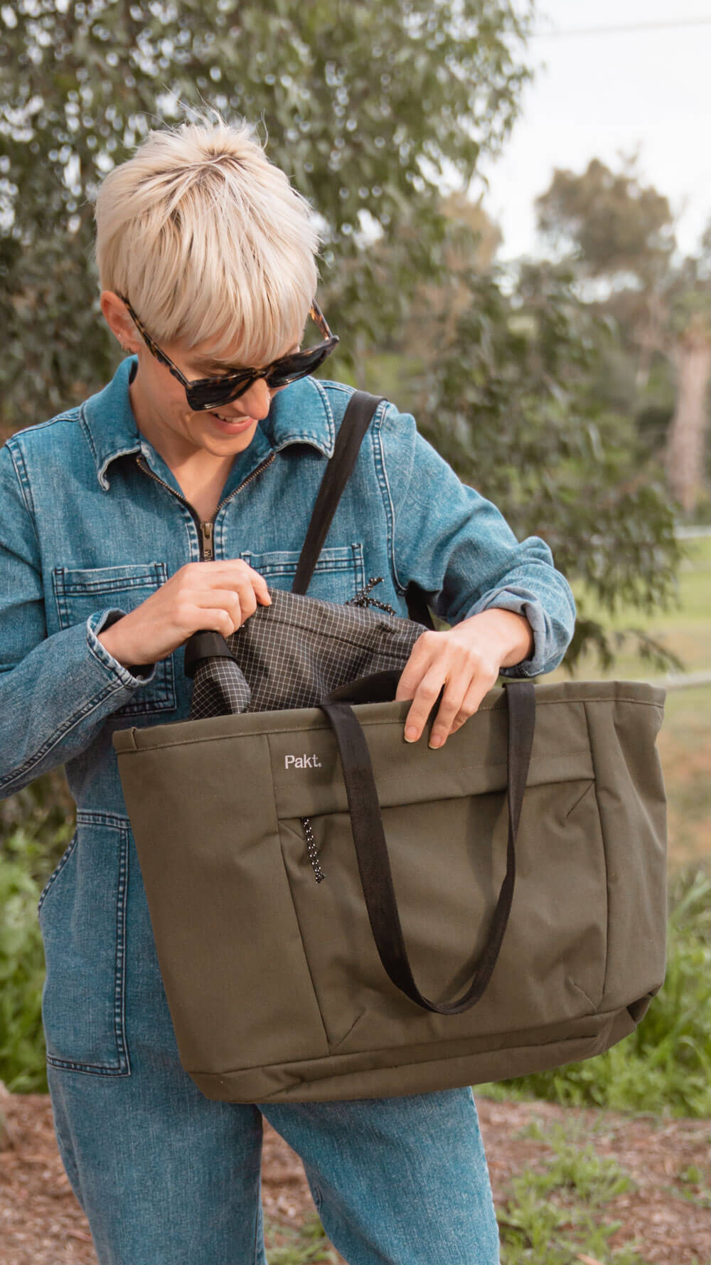Person reaching into cypress Everyday 25L Tote while standing in a park-like outdoor setting
