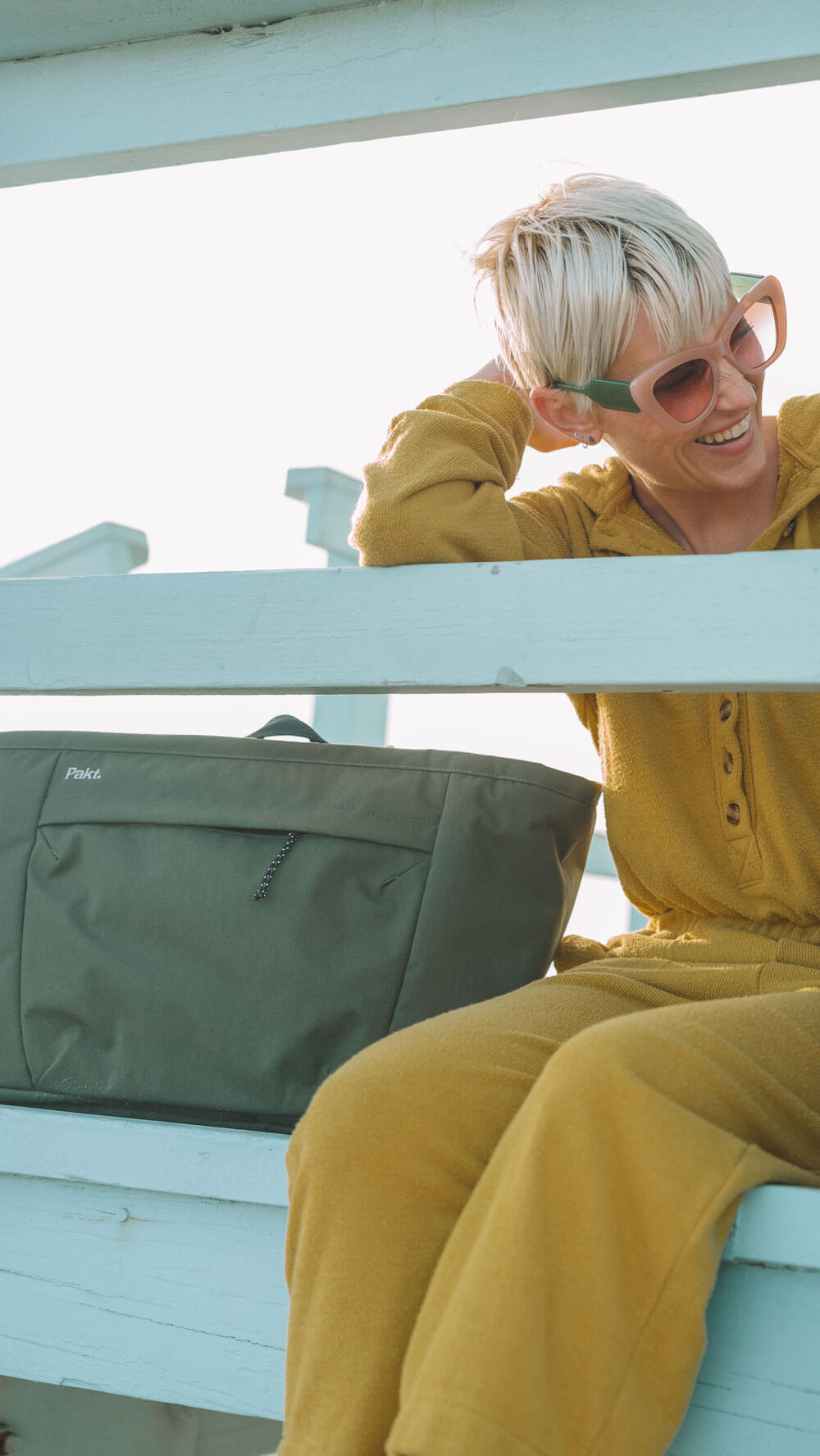 Person with short blonde hair wearing large sunglasses and a mustard yellow outfit sitting on light blue wooden steps next to a green bag labeled 'Pakt.'
