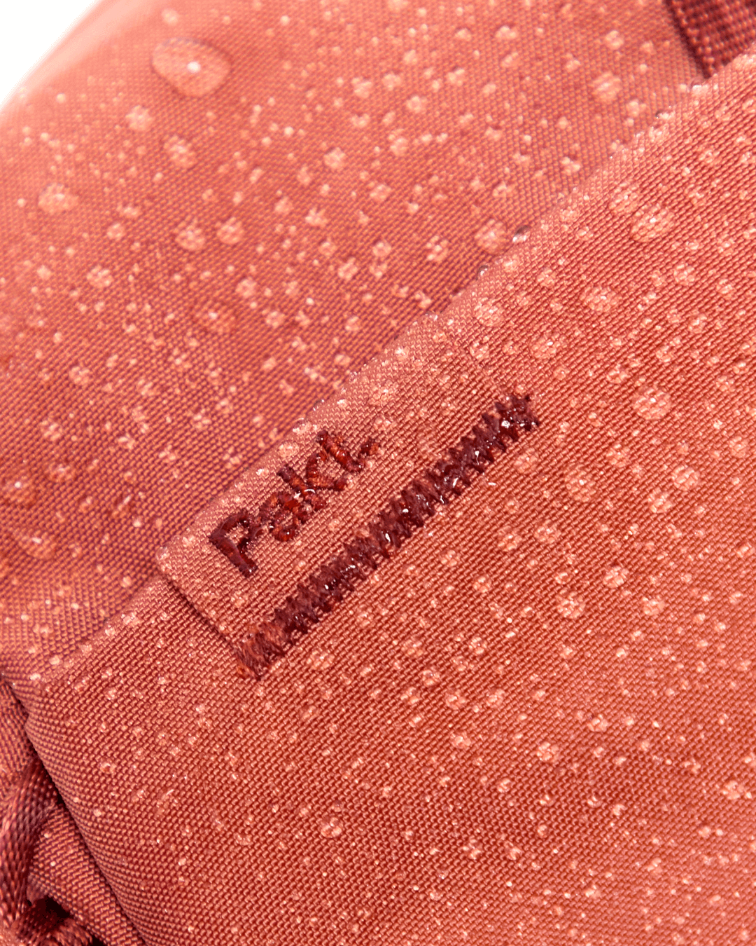 Close-up of MODE Backpack 22L showcasing water beading on water-resistant fabric with a stitched Pakt logo tab in sumac