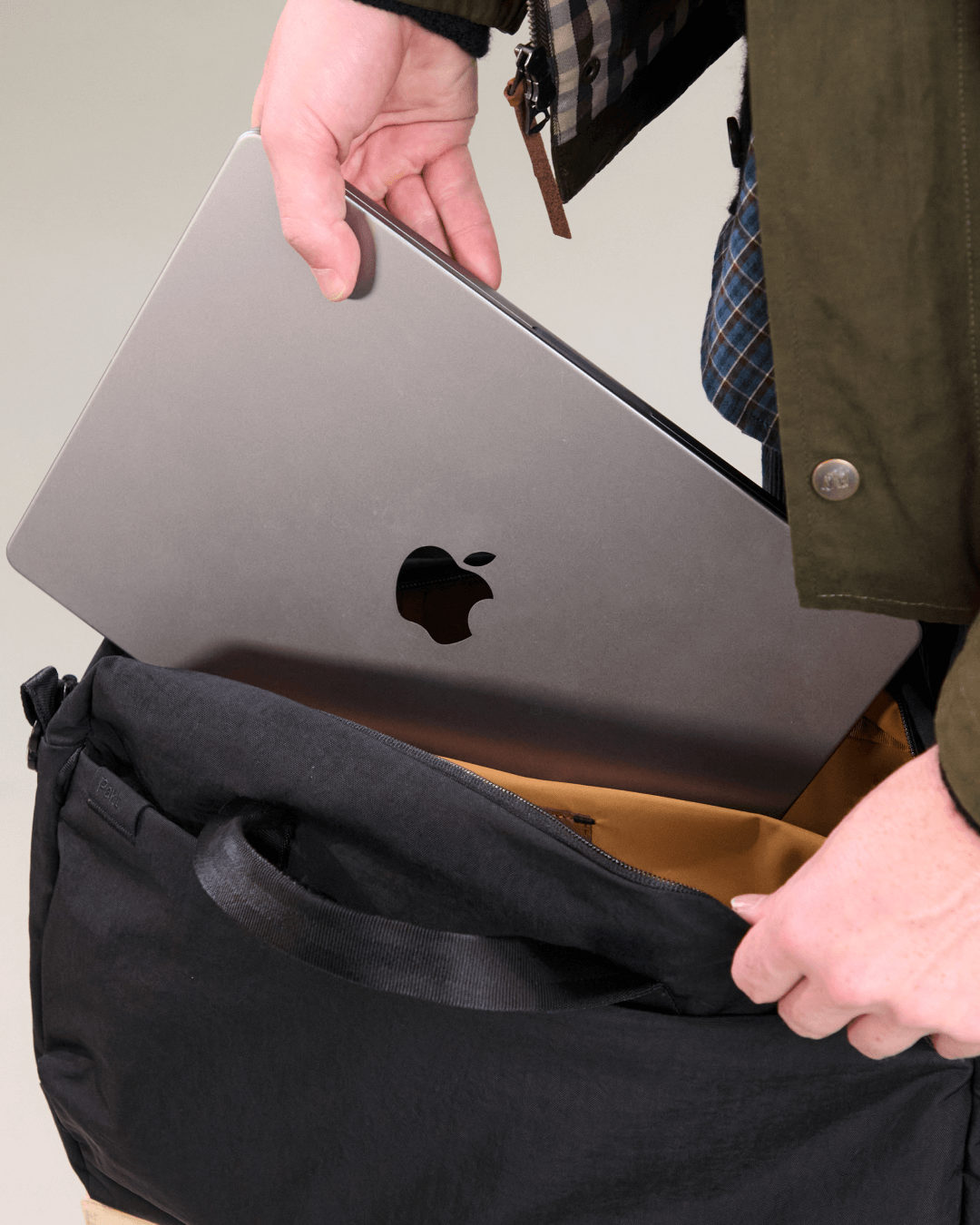 Top-down view of MODE Shoulder Bag 12L as a hand inserts a 16-inch laptop, showing padded laptop sleeve and tan interior