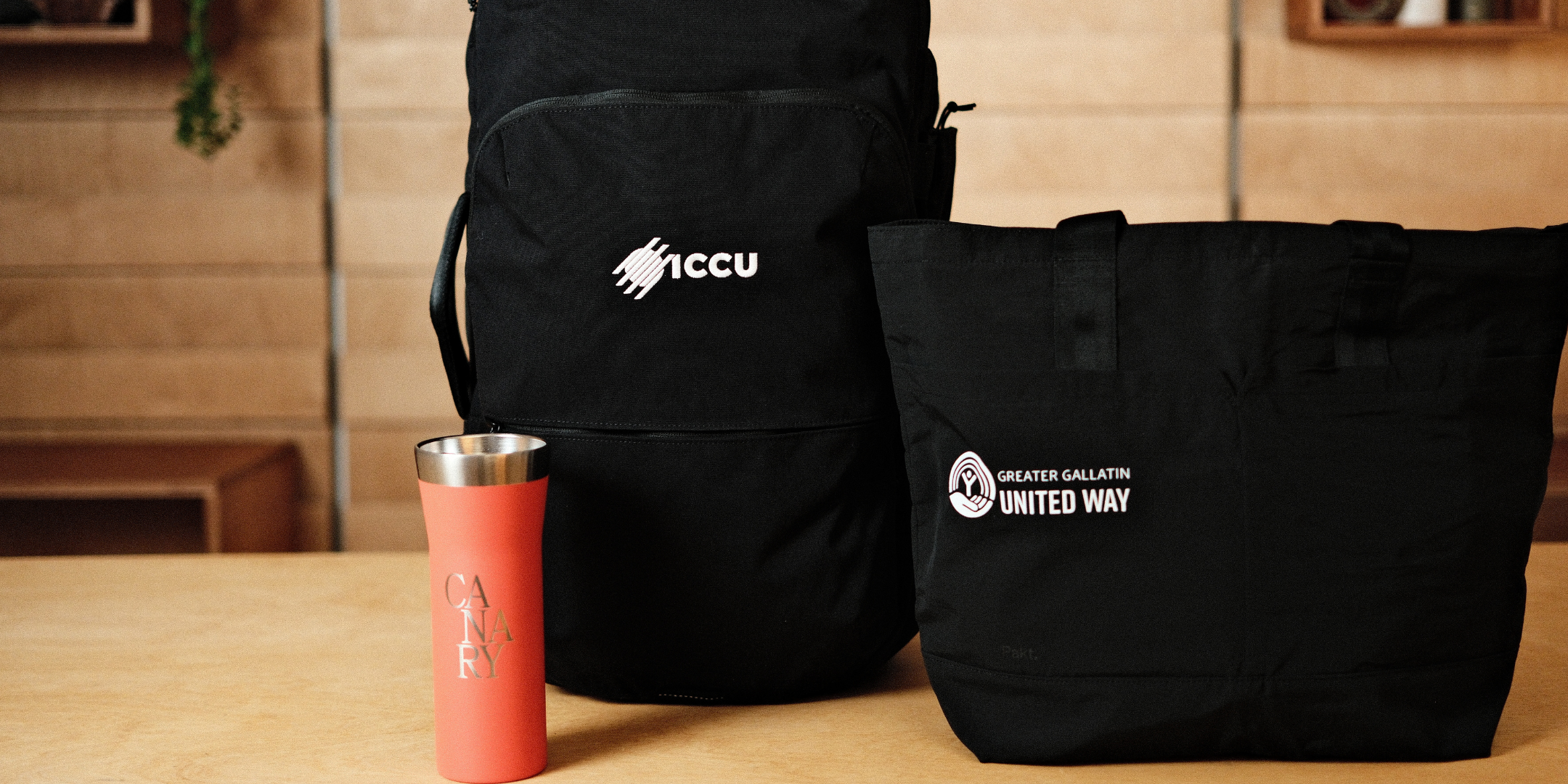 Backpack, branded black tote, and coral travel mug on wood table against wood-paneled wall; tote reads 'United Way', backpack 'ICCU', mug 'CANARY'