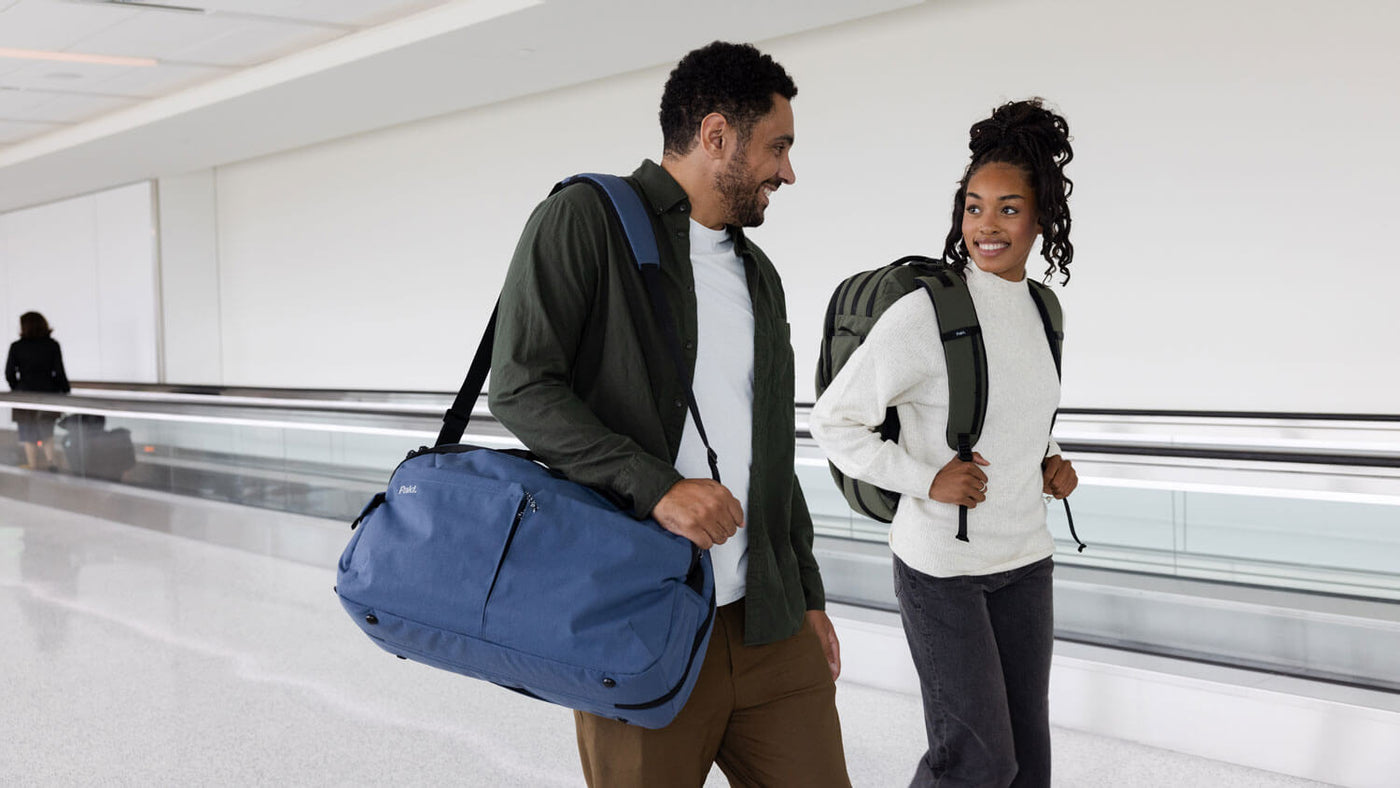 Two people walking in an airport terminal, one carrying a blue duffel bag and the other wearing a green backpack, smiling at each other.