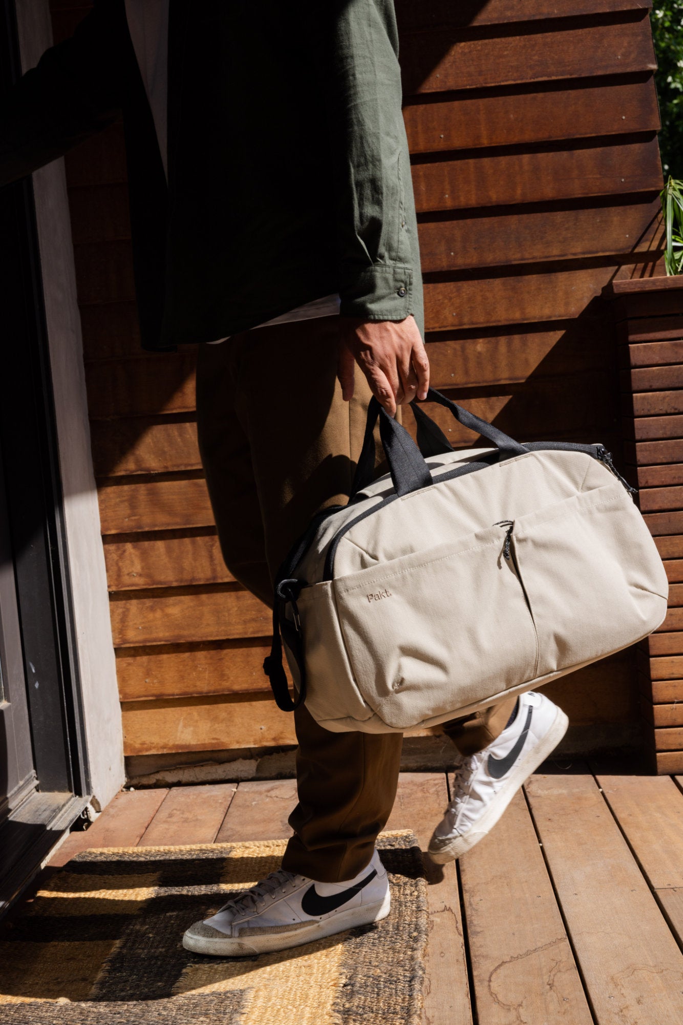 Person holding a 25L Travel Duffel in Sand with black handles labeled 'Pakt', wearing brown pants and white Nike sneakers, standing on a wooden porch.