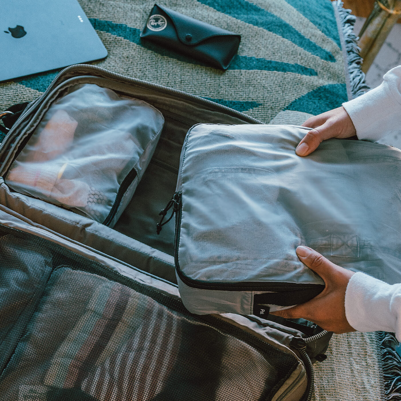 Hands placing a gray packing cube into an open suitcase containing other packing cubes and a black Ray-Ban glasses case on a patterned surface