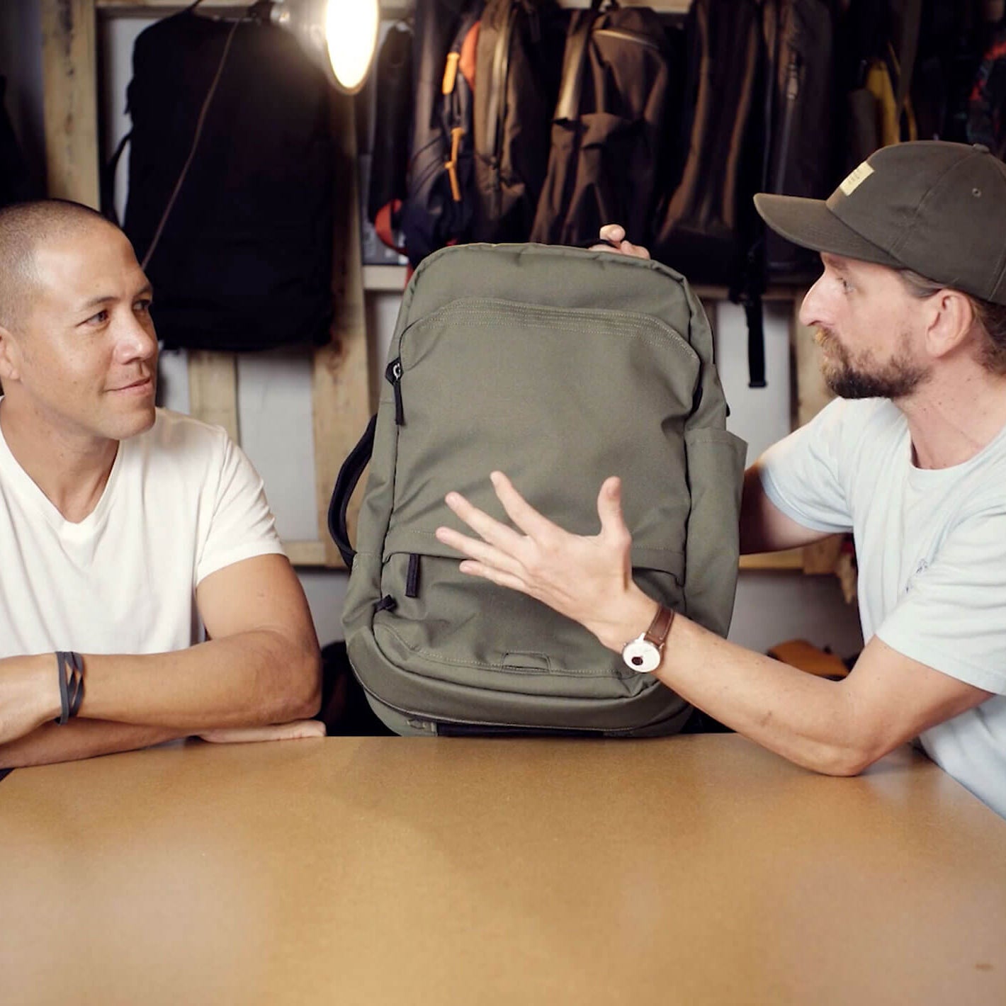 Two men sitting at a table, one holding and showing a green V1 Travel Backpack to the other in a store or workshop setting with backpacks hanging in the background.