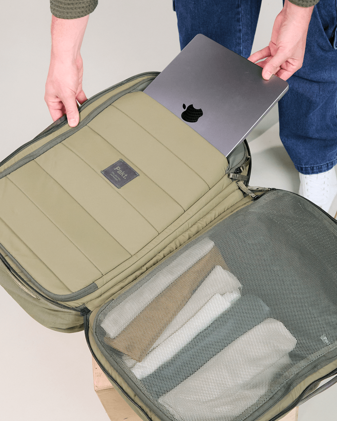 Person placing laptop into olive-green Pakt backpack's padded sleeve; folded clothes visible through mesh pocket.