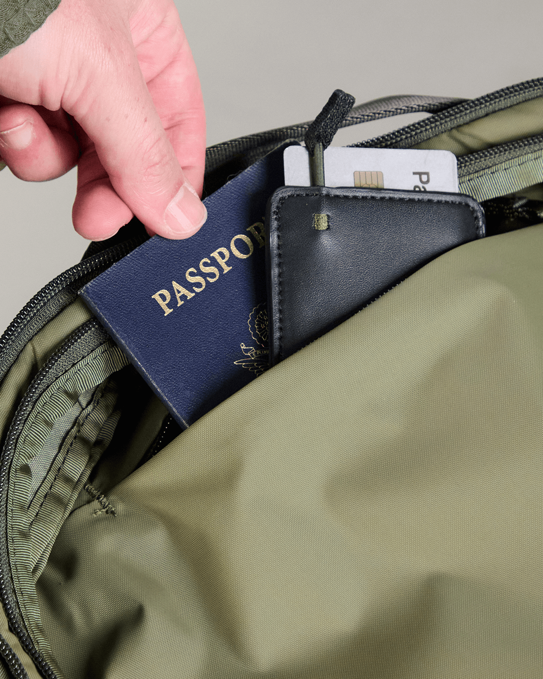 Hand placing a blue passport labeled PASSPORT and a card reading Pa into an olive-green backpack pocket.