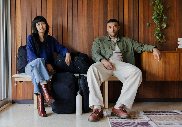 Two people sitting on a bench with travel bags and a water bottle against a wooden wall.