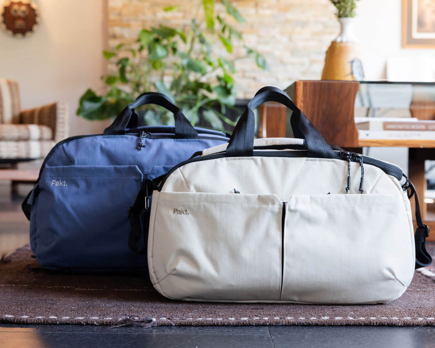 Two Pakt brand bags, one sand and one ocean, with black handles, placed on a rug in a cozy living room setting.