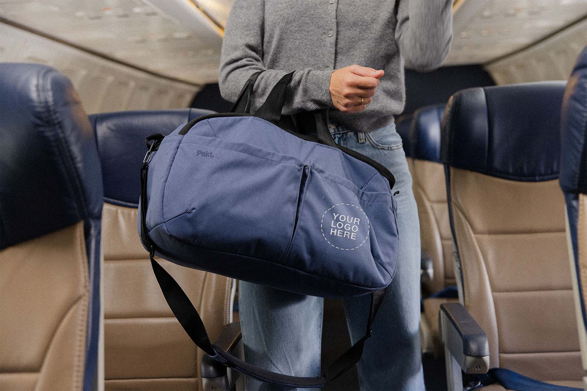 Person holding navy duffel bag with circle text 'YOUR LOGO HERE' and small 'Pakt.' logo in airplane cabin.