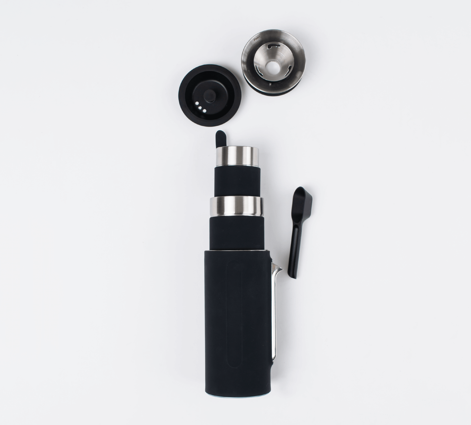 Black Pakt Coffee Kit with a cylindrical main body, a detachable lid, a metal filter, and a small black scoop, all arranged on a white background.