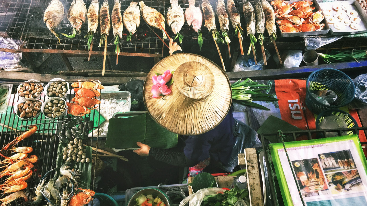 Overhead view of a person wearing a conical hat with pink flowers, preparing grilled fish and seafood at a market stall.