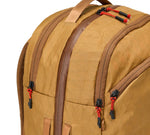 Tan backpack corner showing two zippered compartments with brown trim and red zipper pulls