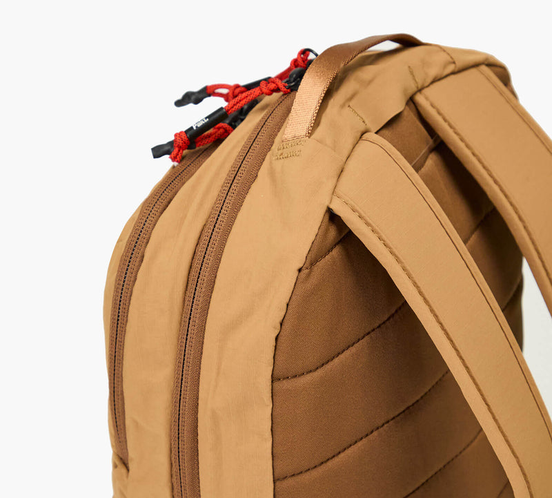 Tan backpack rear showing padded shoulder straps, double zippered compartment and red corded zipper pulls.