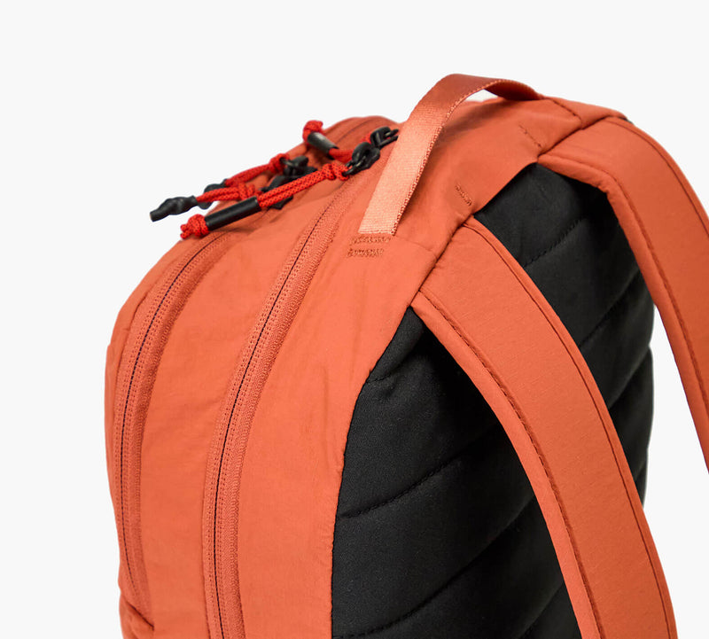 Close-up of orange backpack top: padded black back panel, shoulder straps, carry handle, and zipper with red pulls.