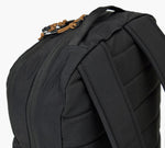 Close-up of black backpack top corner showing padded shoulder strap, zippered compartment, and leather zipper pulls.