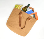 Brown tote bag with handles, containing a wine bottle, popcorn, a blue towel, and a book.