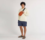 Person wearing a beige long-sleeve shirt, navy shorts, and sandals, modeling an orange Stash Packable Sling 4L bag across the chest.
