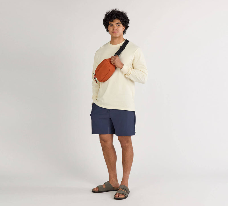 Person wearing a beige long-sleeve shirt, navy shorts, and sandals, modeling an orange Stash Packable Sling 4L bag across the chest.