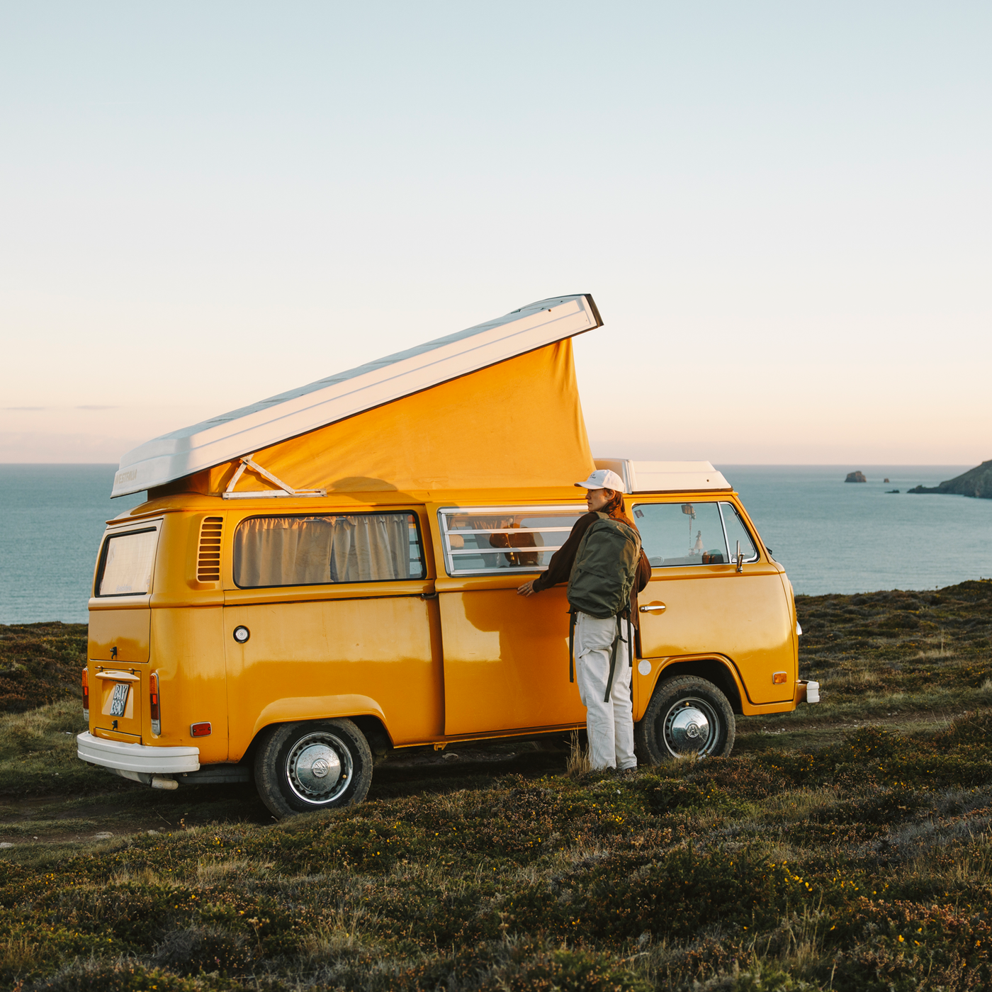 Yellow vintage VW camper van with pop-up roof parked on coastal cliff at sunset; person with backpack standing at side door.