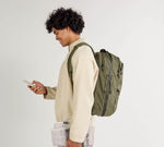 Person with curly hair in beige jacket wearing olive-green backpack, holding smartphone and folded newspaper, side profile.