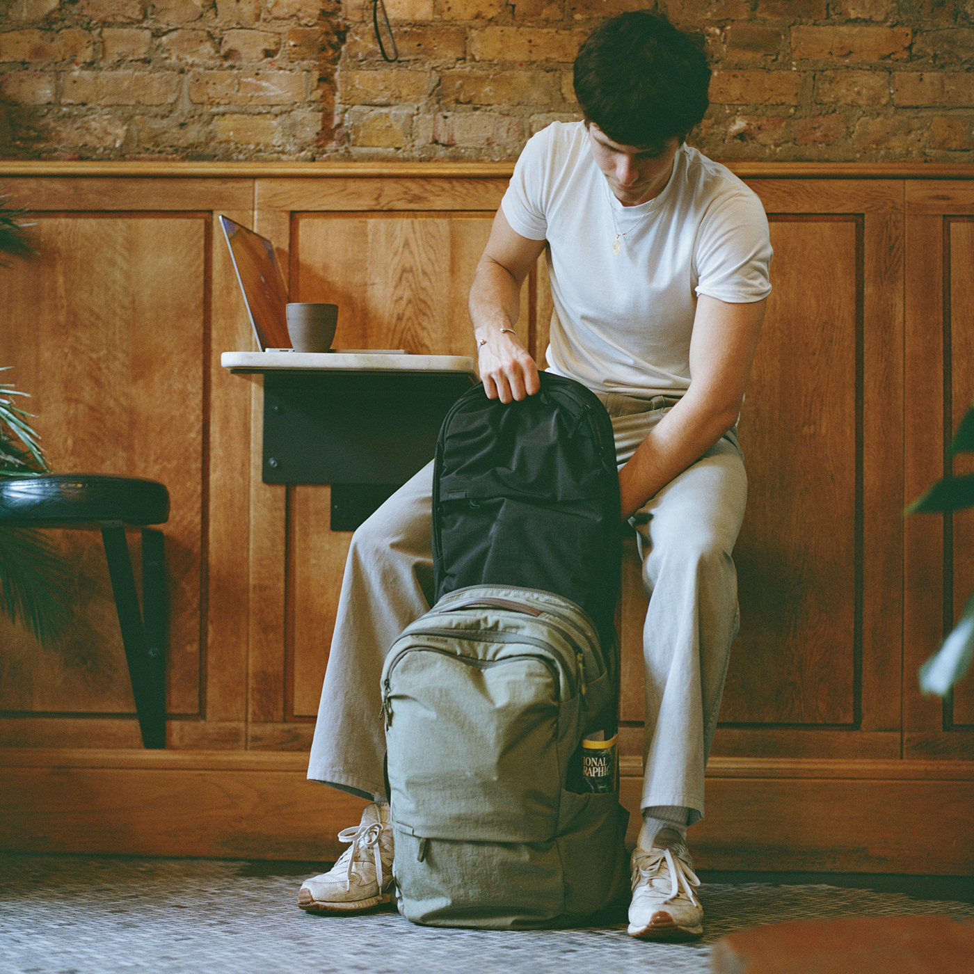 Young man at a cafe table unpacking a backpack beside a laptop and coffee cup