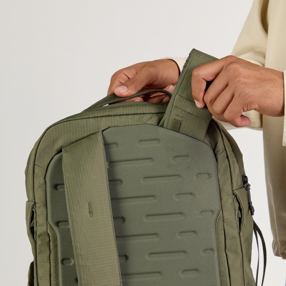 Olive-green backpack back panel and shoulder strap being adjusted by two hands.
