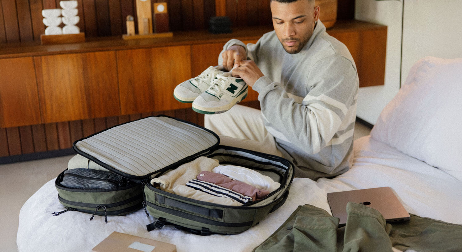Man packing a suitcase on a bed, holding a pair of white and green sneakers, with folded clothes inside the suitcase and a laptop nearby.