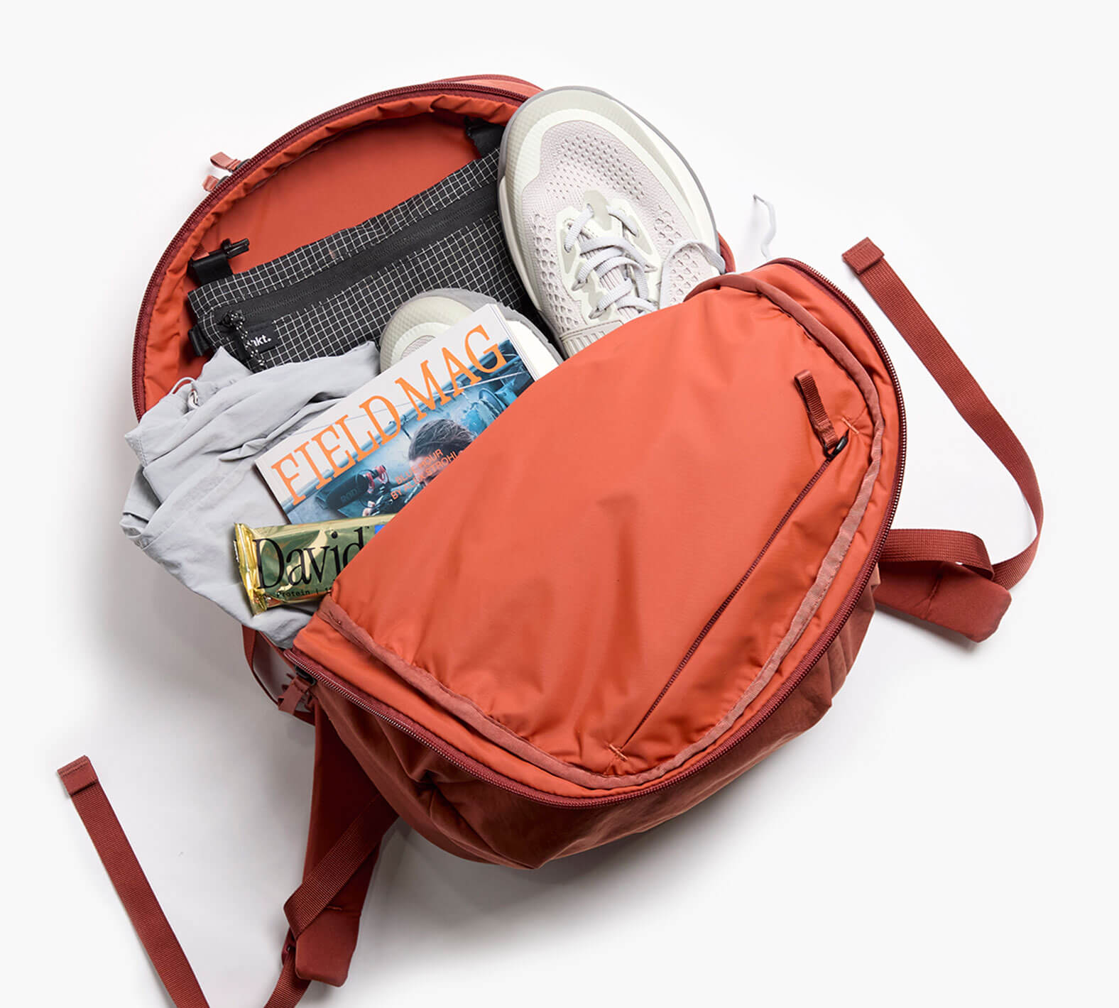 Open rust MODE 22L backpack with sneakers, shirt, FIELD MAG magazine, and David snack bar